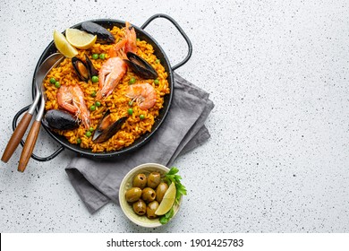 Classic dish of Spain, seafood paella in traditional pan on white wooden background top view. Spanish paella with shrimps, clamps, mussels, green peas, fresh lemon wedges from above, space for text
