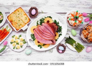 Classic Easter Ham Dinner. Top Down View Table Scene On A White Wood Background. Ham, Scalloped Potatoes, Eggs, Hot Cross Buns, Carrot Cake And Vegetables.