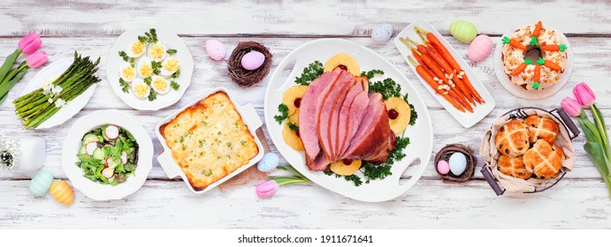 Classic Easter Ham Dinner. Top Down View Table Scene On A White Wood Banner Background. Ham, Scalloped Potatoes, Eggs, Hot Cross Buns, Carrot Cake And Vegetables.