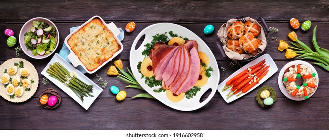 Classic Easter ham dinner. Top view table scene on a dark wood banner background. Ham, scalloped potatoes, eggs, hot cross buns, carrot cake and vegetables.