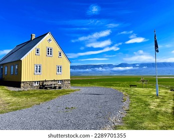 Classic Icelandic wooden yellow house with a rocky base and white windows, accompanied by rocky road and flagpole standind in a lovely green countryside with mountains in the background - Powered by Shutterstock