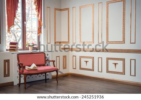 Classic interior design. Elegant interior in red and golden colors with a barocco couch with an open book standing against the window decorated with beautiful curtains and books on the window sill