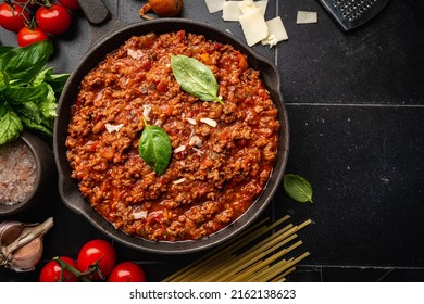 classic italian bolognese sauce stewed in a pan with ingredients on black tile background, top view