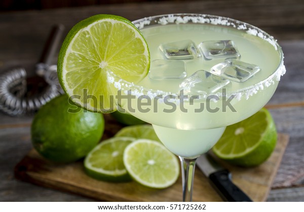 Classic lime margarita cocktail with\
sliced and whole limes sitting on wooden cutting\
board