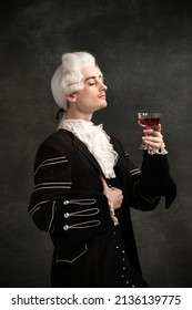 Classic music and wine. Young man wearing wig and vintage medieval outfit like famous composer isolated on dark green vintage background. Retro style, fashion, art, comparison of eras concept.
