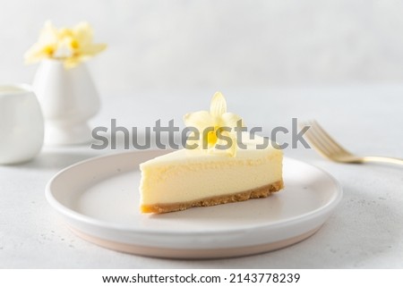 Classic New York cheesecake with fresh vanilla flower on a white concrete background, side view. A piece of Vanilla cheesecake on a white plate. Confectionery menu, recipe. Close up.