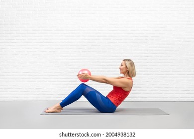 Classic pilates, half roll down drill. Fit woman does practice with pink mini-ball in hands, indoor in loft studio.