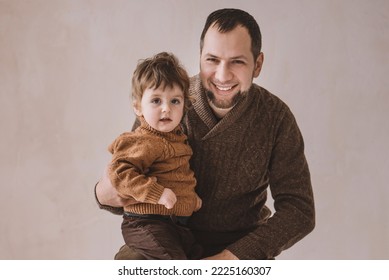 Classic Portrait Of Dad And Son 2 Years Old Portrait On A Beige Background In Coarse Knit Sweaters. Child Sitting On Father's Lap