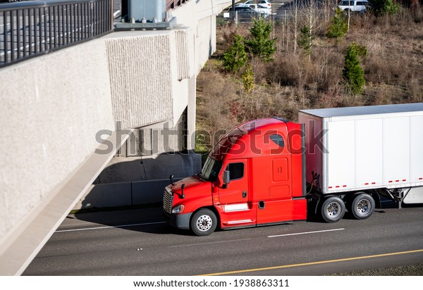 Classic powerful big rig red semi truck tractor\
with day cab and roof spoiler transporting commercial  cargo in dry\
van semi trailer moving on the highway fork intersection with\
bridge across