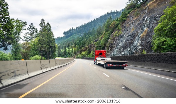 Classic red powerful big rig industrial semi truck\
tractor with day cab transporting empty step down semi trailer\
running on the divided mountain highway road with rock wall and\
trees on the side
