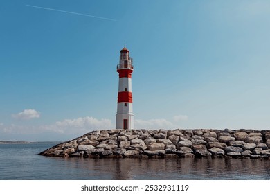 A classic red and white striped lighthouse stands tall on a rocky coast, surrounded by calm ocean waters and a blue sky with fluffy clouds. This maritime landmark offers a sense of peace and tranquili - Powered by Shutterstock