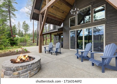 Classic Rustic New Home In Dark Grey Wood Exterior With Forest Landscape And Back Porch With Pire Pit And Four Chairs.