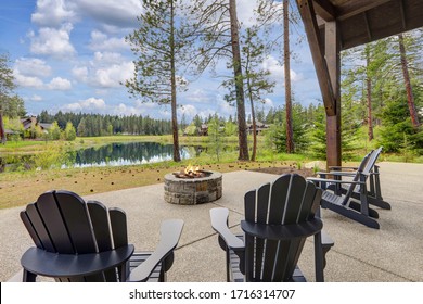 Classic Rustic New Home In Dark Grey Wood Exterior With Forest Landscape And Back Porch With Pire Pit And Four Chairs.