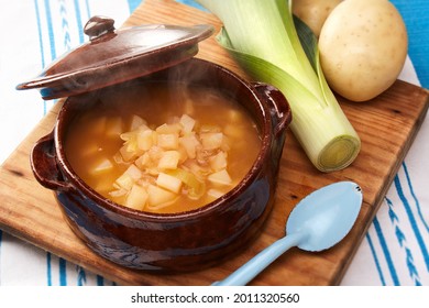 Classic rustic potato and leek soup served in a clay or earthenware bowl over a wooden cutting board by a leek, a couple of potatoes and an old blue spoon. Traditional cooking. - Shutterstock ID 2011320560
