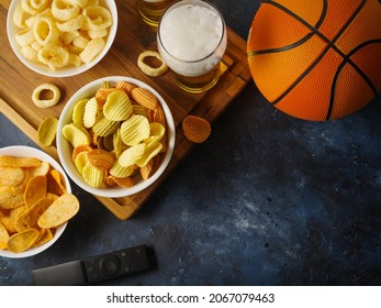 Classic set of sports fans - light beer, cold snacks on a wooden tray. TV remote and basketball on the table. Blue background. A high angle view. Sports on TV, rest in the company of friends.