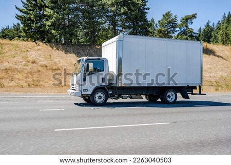 Classic small white cab over rig semi truck tractor with box trailer for local deliveries transporting commercial cargo driving on the wide highway road with trees on the hill