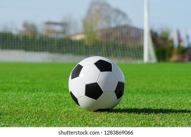 Classic Soccer Ball Lies On The Bright Green Grass On The Football Field Against The Background Of The Stands For Fans And The Football Goal At A Sports Stadium Close-up In A Large Sports Center