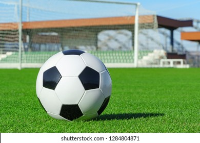 Classic Soccer Ball Lies On The Bright Green Grass On The Football Field Against The Background Of The Stands For Fans And The Football Goal At A Sports Stadium Close-up In A Large Sports Center