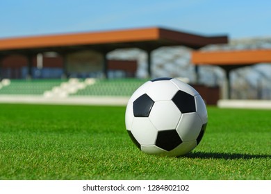 Classic Soccer Ball Lying On The Bright Green Grass On The Football Field In The Background Of The Stands For The Fans At The Sports Stadium Close-up In A Large Sports Center For Football Players	
