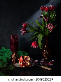 Classic still life with beautiful red tulip flowers and candle, pomegranate, green napkins that appeared in a vintage vase. Art photography. Front view