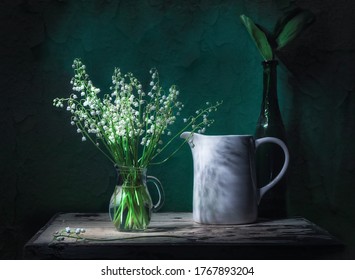 Classic still life with beautiful white Lily of the valley bouquet in glass jar and a white jar in ray of light. Green background. Art photography.