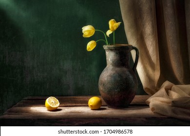 Classic still life with bouquet of three yellow tulip flowers in old vintage jug, two cut lemon fruits and drapery in beam of light on green background and old wooden table. Art photography.