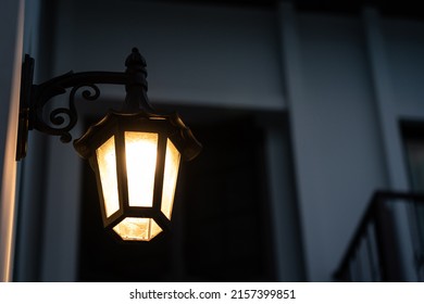 A Classic Style Street Lighting Lamp Is Glowing In Orange Warm Light Shade In Evening Night Time With Background Of Building Structure. Photo Contained Noise Due To Dark Condition. Selective Focus.