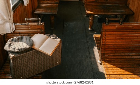 A classic suitcase, flat cap, and open book rest on a wooden bench inside an old-fashioned train carriage, bathed in sunlight