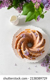 Classic Vanilla Bundt Cake With Sugar Glaze And Lilac Flowers On White Background. Spring Composition. Homemade Lemon Butter Bundt Cake Recipe. Bakery, Confectionery Menu. Top View, Flat Lay