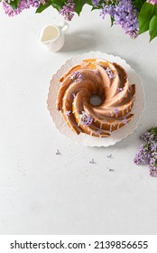 Classic Vanilla Bundt Cake With Sugar Glaze And Lilac Flowers On A White Background. Spring Composition. Homemade Lemon Butter Bundt Cake Recipe. Bakery, Confectionery Menu. Top View, Copy Space