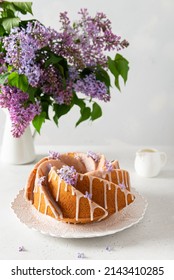 Classic Vanilla Bundt Cake With Sugar Glaze And Lilac Flowers On A White Background. Spring Composition. Homemade Lemon Butter Bundt Cake Recipe. Bakery, Confectionery Menu. Side View, Copy Space