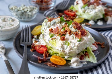 Classic wedge ice berg lettuce salad with blue cheese dressing, bacon, tomatoes, onions, chives sitting on gray plates with ingredients on the side. Keto.