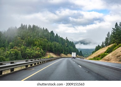 Classic white big rig semi truck with high cabin for long haul freight transporting commercial cargo in dry van semi trailer running on the winding divided mountain road with trees on the hillside