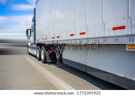 Classic white powerful long haul diesel big rig semi truck with high cab transporting cargo in dry van semi trailer with aerodynamic skirt running on the wide straight road to point of delivery