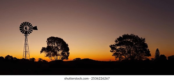 A classic windmill stands tall against a vibrant sunset sky, silhouetted by the surrounding trees and rolling hills. The warm hues of the sky create a sense of tranquility and nostalgia - Powered by Shutterstock