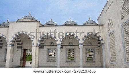 A classical mosque portico featuring white marble columns and striped arches. Decorative stone windows add detail to the structure. The setting is bright and clear.