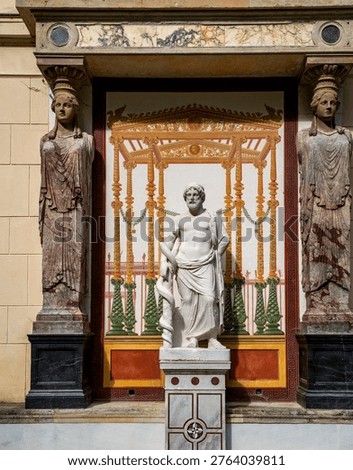 Classical museum display with marble statue of a draped male figure between two caryatid columns and a colorful ancient fresco backdrop, evoking greek and roman art and architecture.