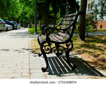 Classical shape old wooden and cast iron park bench along concrete paved sidewalk. diminishing perspective. lush green spring foliage and trees beyond. parks and outdoor. strong shadow. parked cars. - Powered by Shutterstock