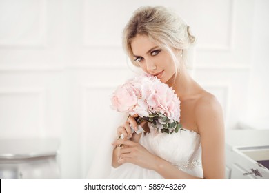 Classical Young Gourgeous Bride. Studio Interior Fashion Shot Of Fashion Model In Wedding Dress With Bouquet Of Flowers Sitting In White Room. Blonde Woman Portait With Profeshional Make-up And