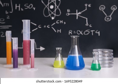 Classroom Desk With Flasks With Colored Chemicals, Instruments And A Drawn Blackboard With Science Teaching In The Background, Chemical Sciences Education Concept