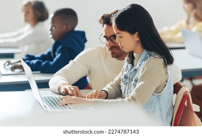 In a classroom full of kids, a male tutor helps a young student with a coding as part of a computer science lesson. Digital literacy teacher providing individual guidance to young minds in a school. - Powered by Shutterstock