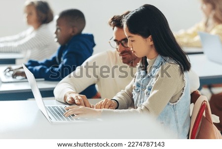 In a classroom full of kids, a male tutor helps a young student with a coding as part of a computer science lesson. Digital literacy teacher providing individual guidance to young minds in a school.