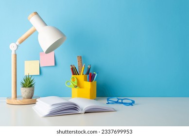 Classroom setting: Side view photo of school supplies neatly placed on desk. Pens, ruler, open book, sticky notes, lamp, glasses add to arrangement. Blue wall backdrop allows for text or ad insertion - Powered by Shutterstock