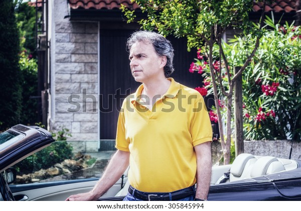 Classy 40 years old sportsman with three-day\
beard and salt and pepper hair wearing a yellow polo shirt while he\
is sitting on the door of a dark brown cabriolet car in residential\
neighborhood