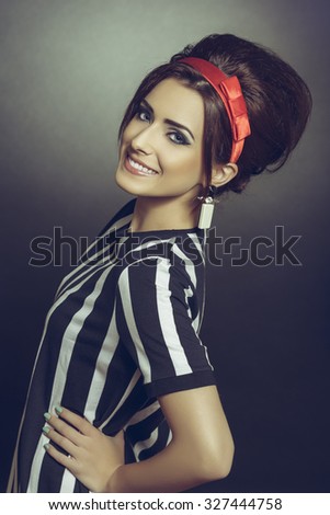 Classy brunette young woman in retro black-and-white stripped attire, accessorized with red headband and 60s hairdo, smiling confident and posing with hand on hip against dark background.