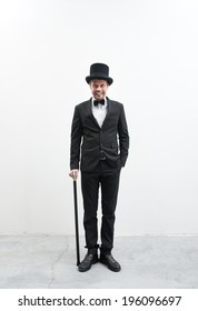 Classy Smiling Gentleman Standing On White Background And Concrete Floor In Elegant Suit With Cane And Bowler Hat.