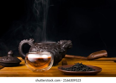 Clay teapot with red tea Lapsang souchong on a black background, heap of tea and glass thermo cup of hot tea with vapour.