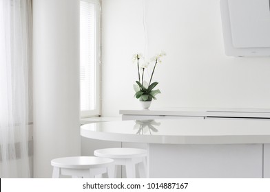 A Clean, All White Kitchen Interior. Scandinavian Light And Modern Home Office In Finland, View From The Living Room. A White Orchid Reflecting From The Table Surface