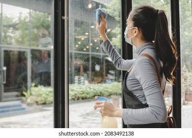 Clean. Asian Waitress Staff Wearing Protection Face Mask In Apron Cleaning Window With Disinfectant Spray Protect Infection Coronavirus (covid-19) In Cafe Coffee Shop Restaurant, Hotel Service Concept