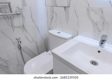 A clean bathroom features white marble-patterned tiles, a white toilet, and a matching sink. The design is minimalist and modern, emphasizing hygiene
